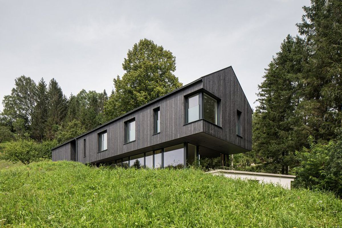 A black gable-roofed house sits on a grassy plot of land. The top level cantilevers over the glassy lower level.
