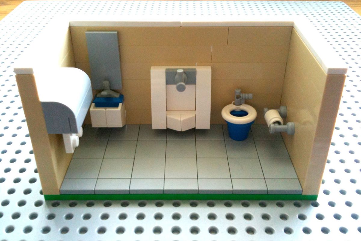Lego model of a public bathroom with light brown walls, toilet, urinal, sink, toilet paper holder, paper towel dispenser, greenish floor, on white and green grid