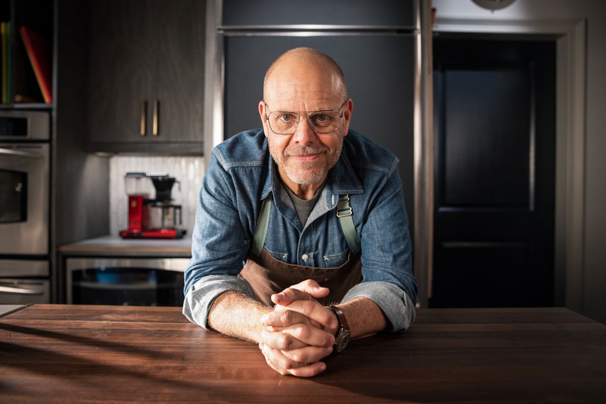 A man wearing a denim shirt and a brown apron leans forward over a wooden countertop.