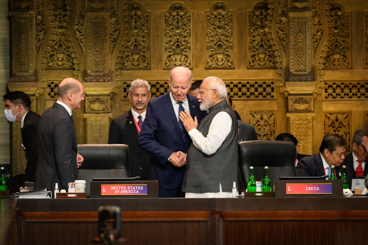 Biden, in the middle of a large room with tables and microphones everywhere, tilts his head down to listen to Modi, on the right, who is speaking to him.