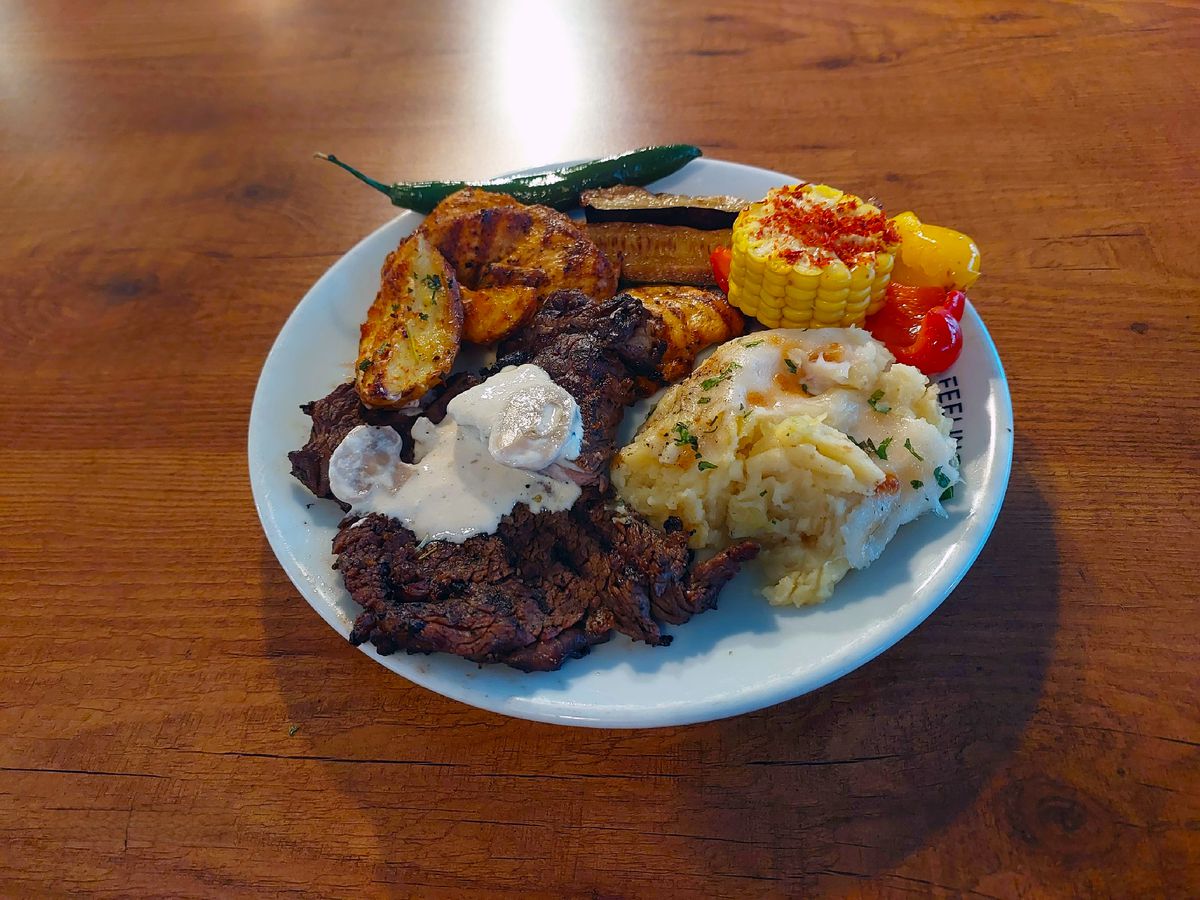A plate of steak topped with a dollop of creamy butter, along with roasted potatoes, zucchini, corn, bell pepper, and mashed potatoes.