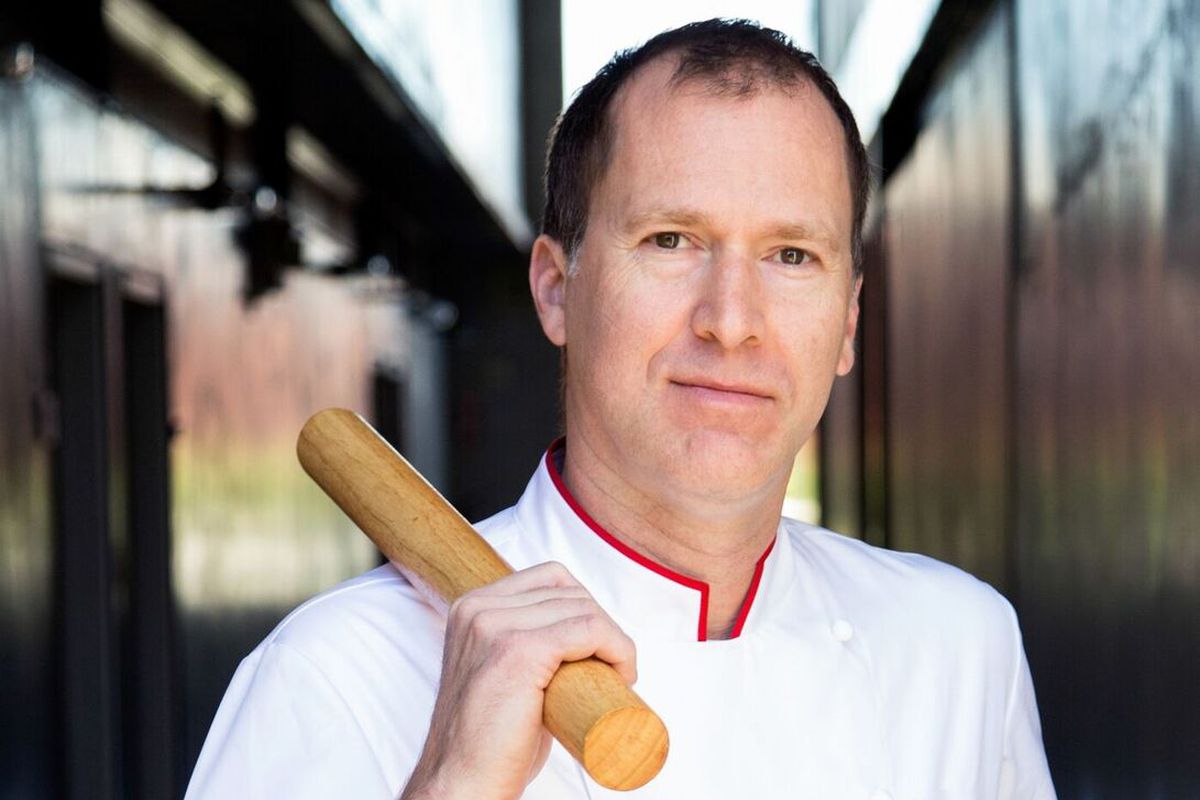 Tall chef Lincoln Carson stands in whites holding a rolling pin.