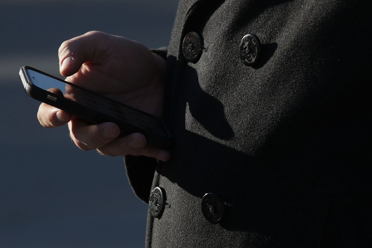 A man in a dark double-breasted coat holds a cellphone and uses his thumb to scroll on the screen.