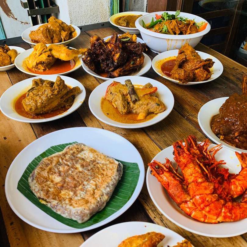 A wooden sunlit table filled with various rich dishes including massive prawns, stuffed flatbread, curries, meaty mains, and a large vegetable salad bowl.