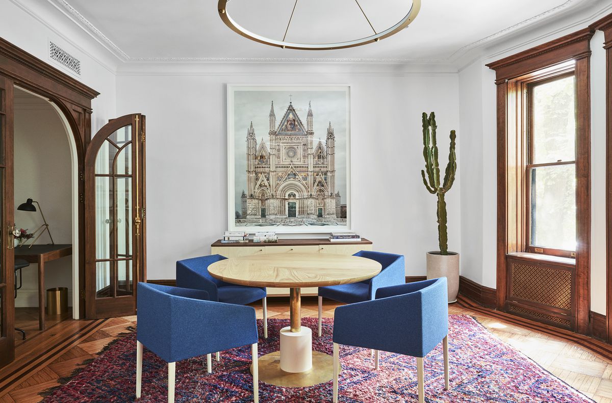 A library. There is a wooden table flanked by multiple blue chairs. The floor is hardwood and there is a patterned area rug under the table. There is a large work of art hanging on the far wall. There is a window letting in natural light. 