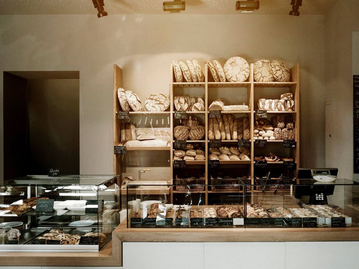 A bakery interior with plain gray walls and exposed beam ceiling. Behind a pastry counter is a large wooden shelf with various kinds of bread