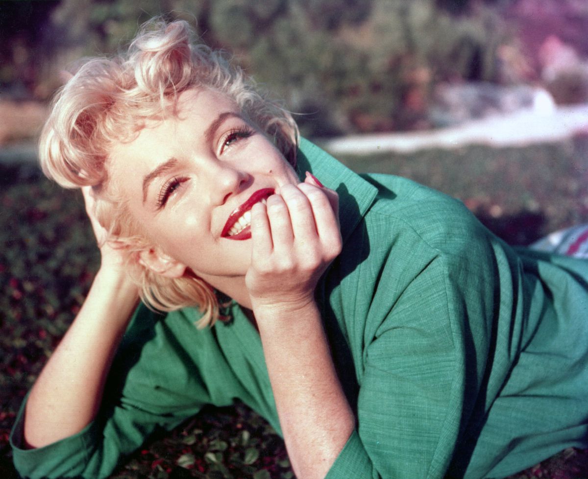 Marilyn wears a green sweater as she lies on her stomach in the grass, propped up on her elbows. Her hair is tousled but her face is immaculately made up, complete with bright red lipstick.