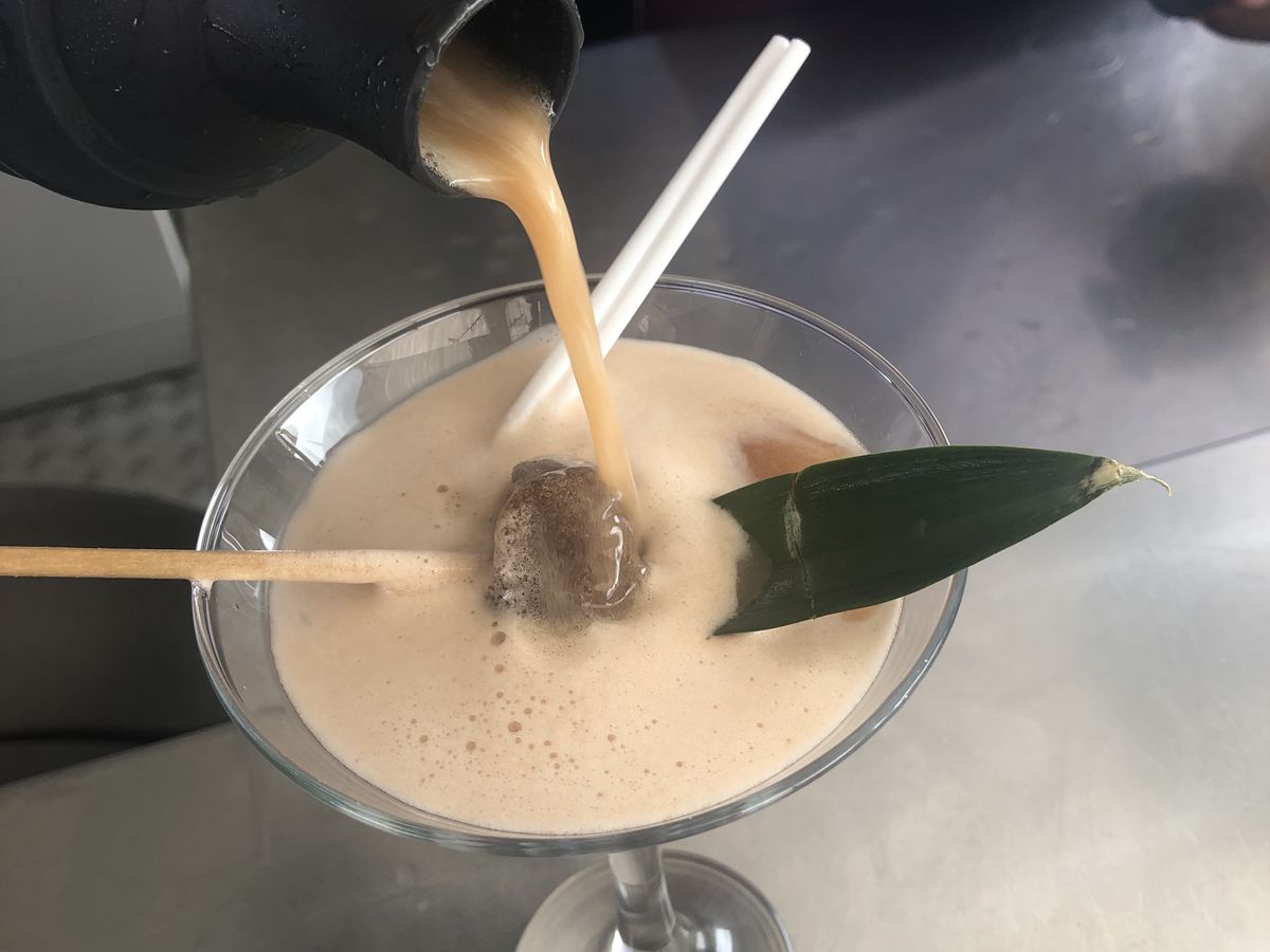 A server pours a thick cocktail from a shaker into a martini glass, garnished with a piece of tamarind candy on a skewer and a tropical leaf