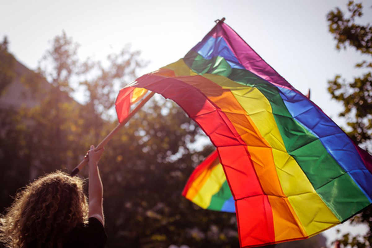 Photo of white woman from back waving a large, backlit Pride (rainbow) flag in city street.