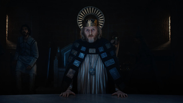 Sean Harris as King Arthur, standing in his court, lit from above, in The Green Knight