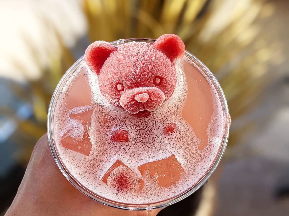 From above, a hand holds a plastic cup with a bright pink drink, garnished with a large floating ice cube in the shape of a teddy bear