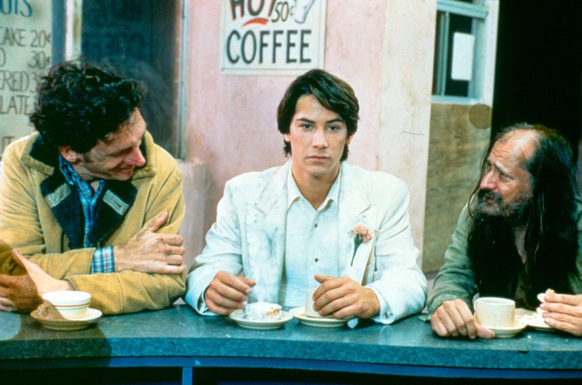 A young Keanu Reeves, wearing a white suit with a pink flower pinned to it, sits at a diner counter between two men talking to him but looks like he isn’t listening.