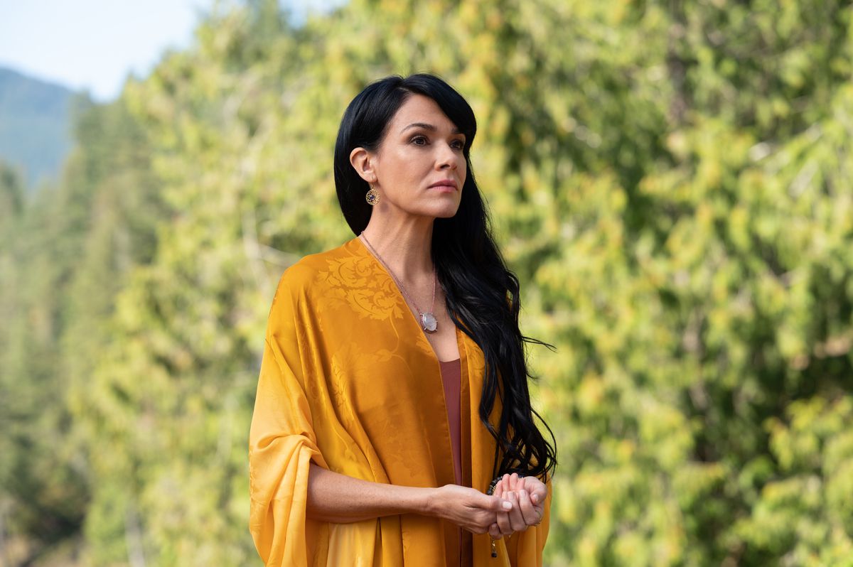 Adult Lottie stands in a picturesque woodsy setting in a flowing yellow shawl in the Yellowjackets season 2 premiere.