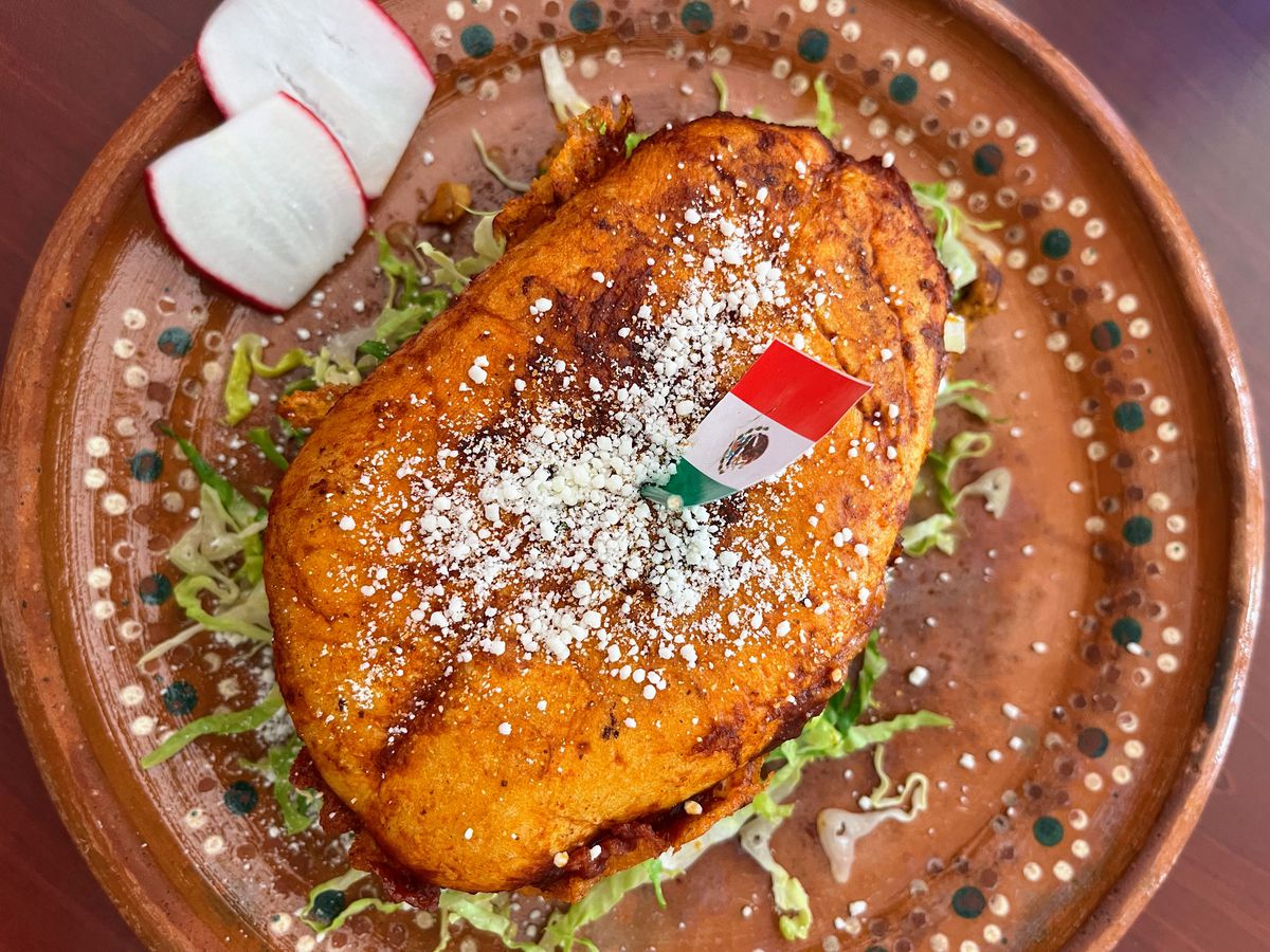 A sandwich with bright orange toasted bread and a sprinkling of white feta on top, with a small Mexican flag stuck in the middle of the sandwich.