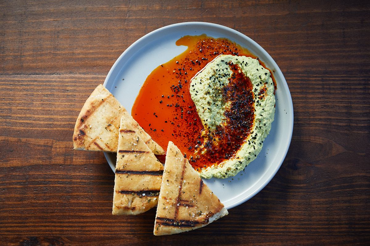 a white plate on a wooden table of hummus with a bright orange swirl and grilled pita bread
