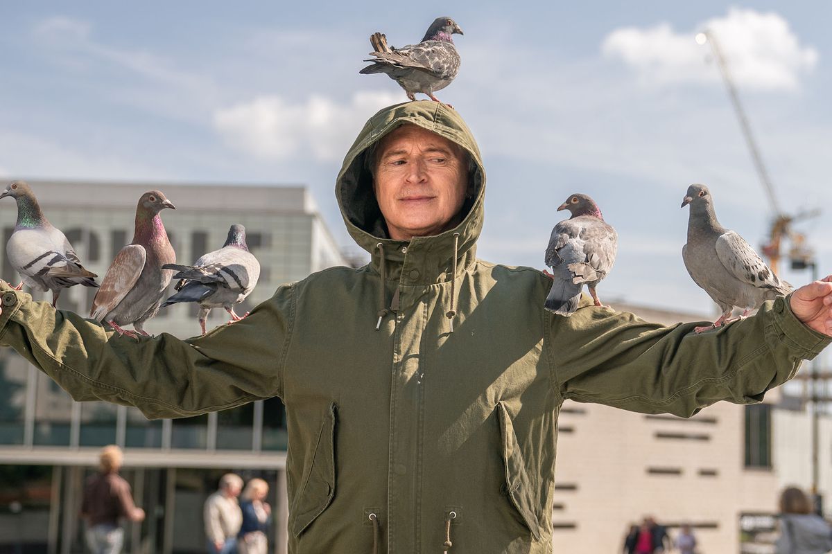Gaz (Robert Carlyle) stands in an open square with buildings all around him, arms extended, wearing a green coat with the hood pulled up, with pigeons perched on his head and arms in a PR image from FX’s TV series The Full Monty