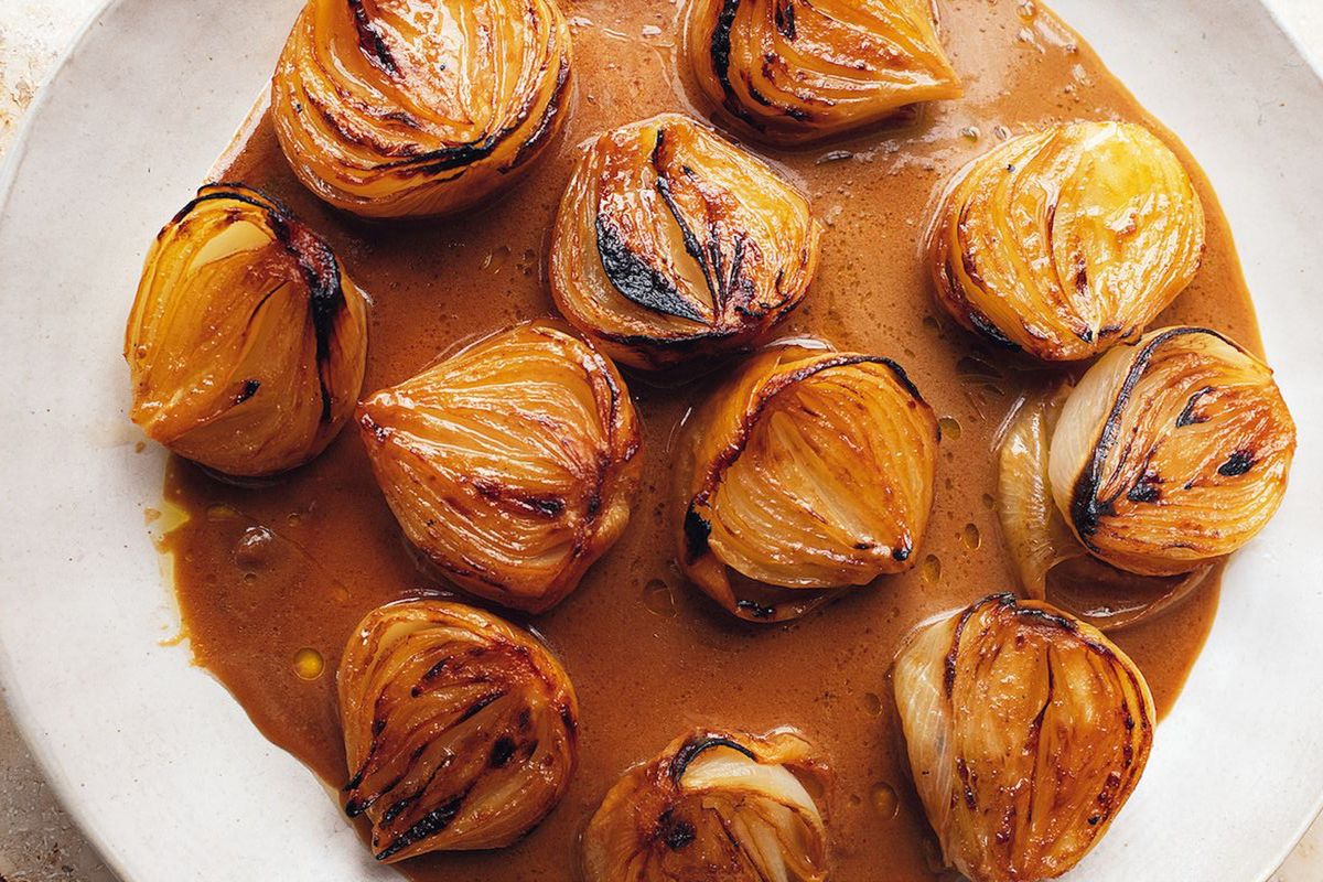 A platter of baked, slightly charred onions in a deep brown miso bitter sauce, served on a large white platter; it’s a recipe from Yotam Ottolenghi, in Flavour