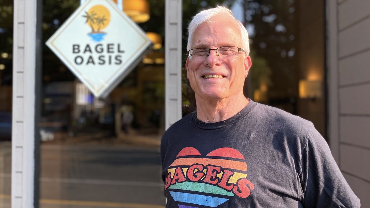 An older white man in glasses and a shirt that read “bagels” stands outside Bagel Oasis