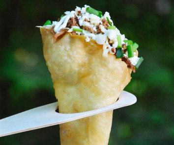 A fried tortilla cone holds some browned pork topped with a white drizzle of sauce and green onion