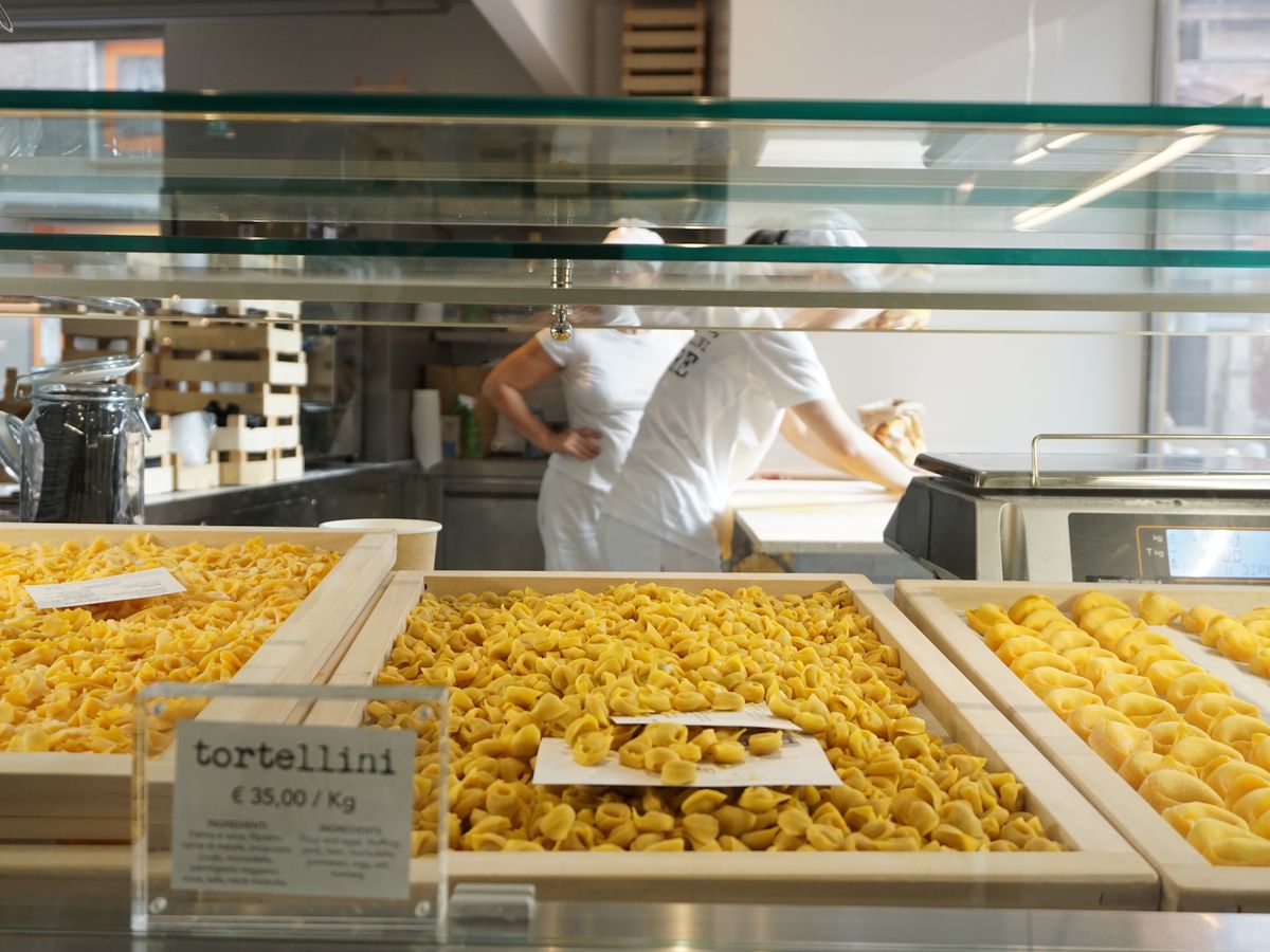 Trays of rolled pasta in a glass case, with kitchen workers beyond