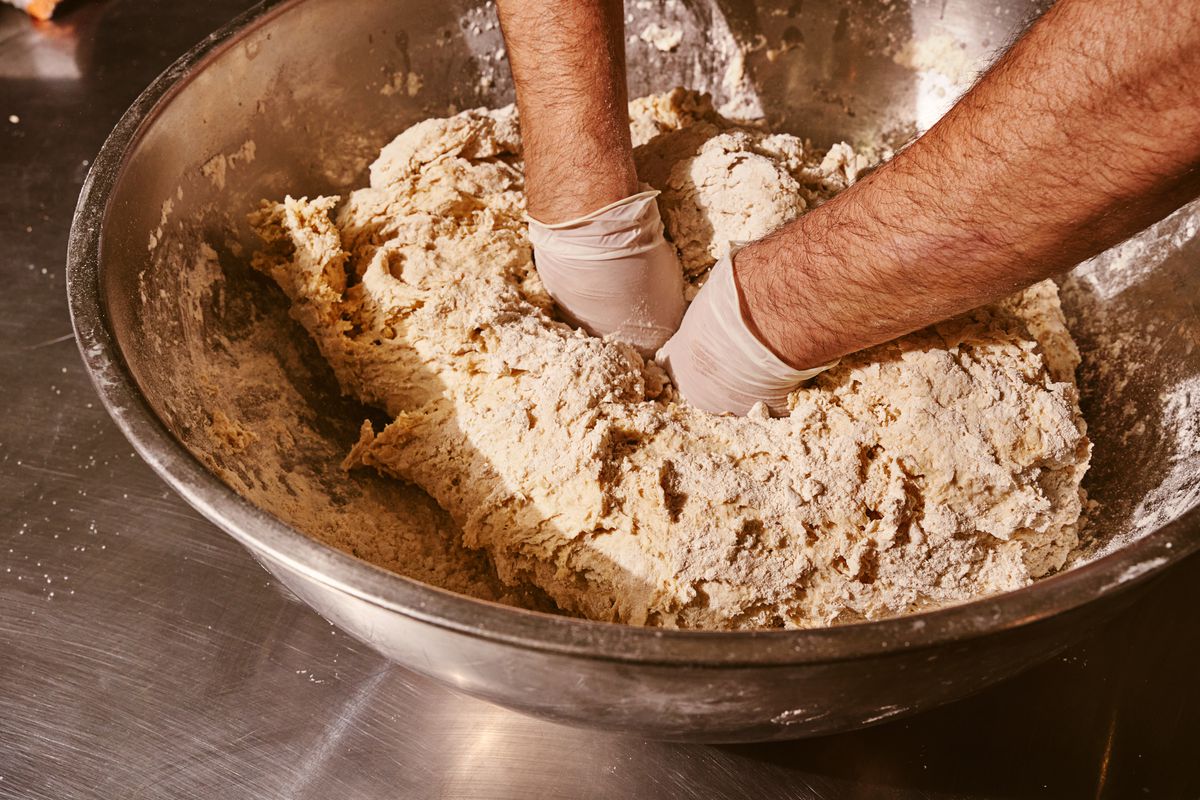 Two gloved hands are buried in a sizable mound of flour tortilla dough in a stainless steel bowl