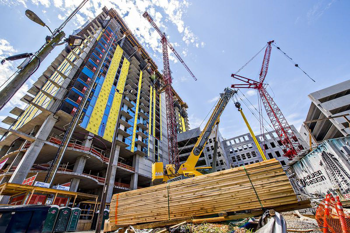 A concrete apartment tower under construction with red cranes and piled wood at the base.
