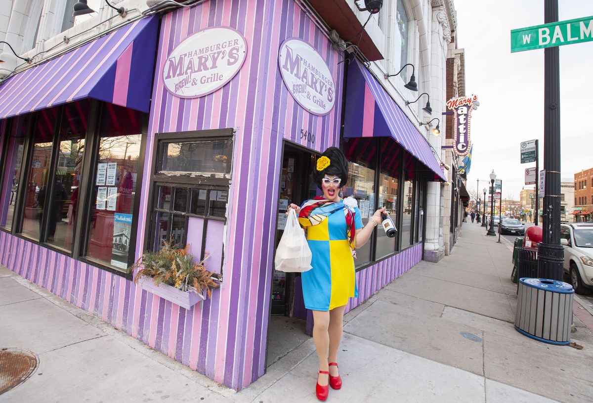 A drag queen stands outside a pink and purple striped corner restaurant.