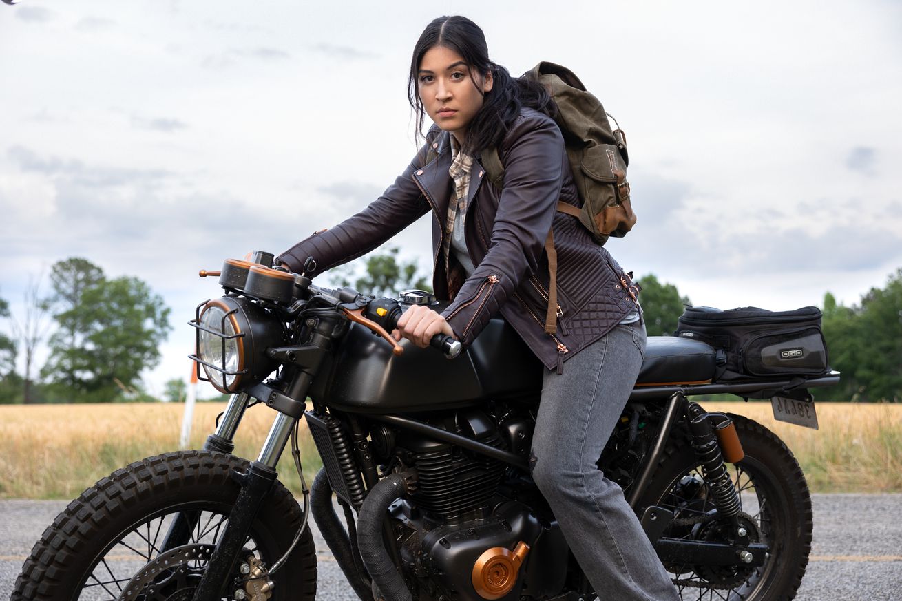 A wide shot of a woman with long black hair tied back in a pony tail, wearing a leather jacket, jeans, and straddling a motor cycle.
