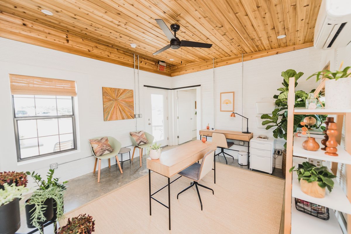 Room with white concrete walls, a wood ceiling, and polished concrete floors. It’s furnished with modern office tables and chairs and has shelves with many plants on them.
