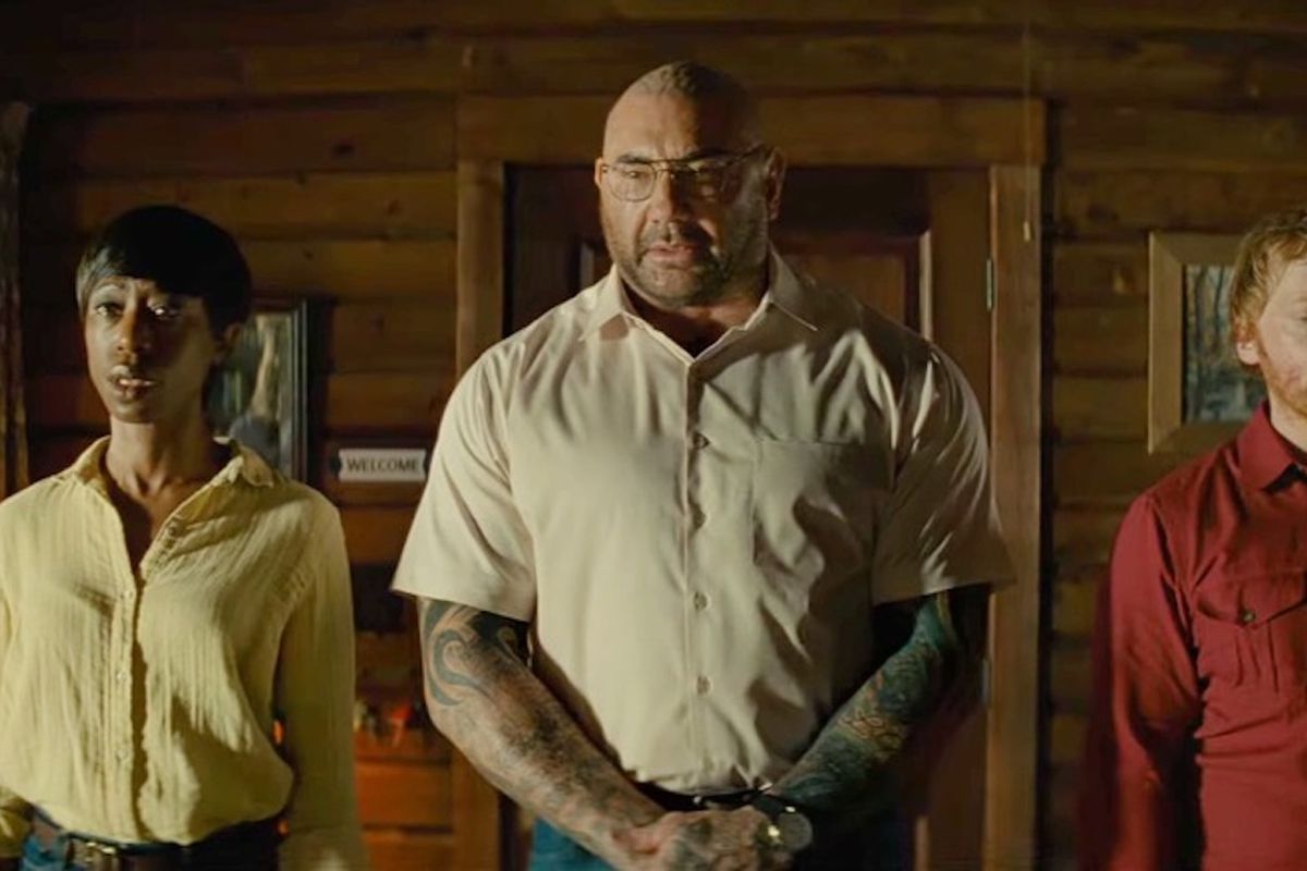 Nikki Amuka-Bird, Dave Bautista, and Rupert Grint stand with stiff posture in the cabin in Knock at the Cabin. They look unnerved and upset.