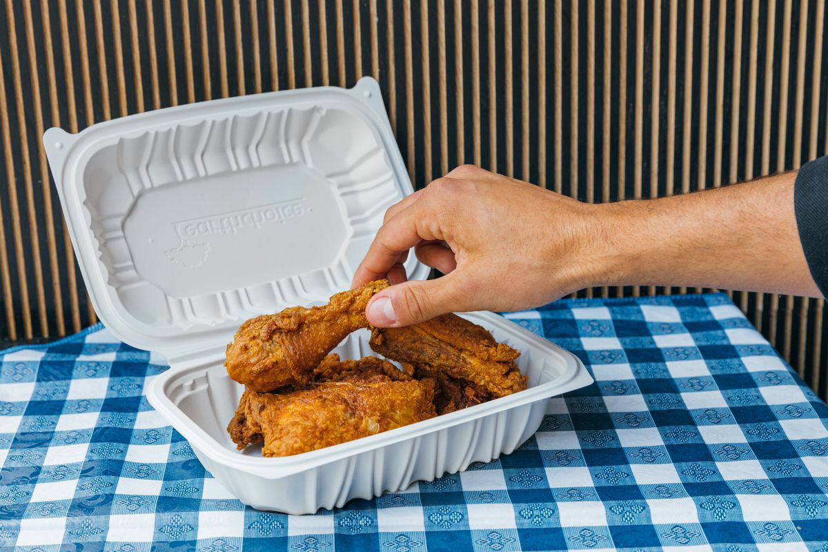 A hand grabs a drumstick from a takeout container of golden fried chicken.