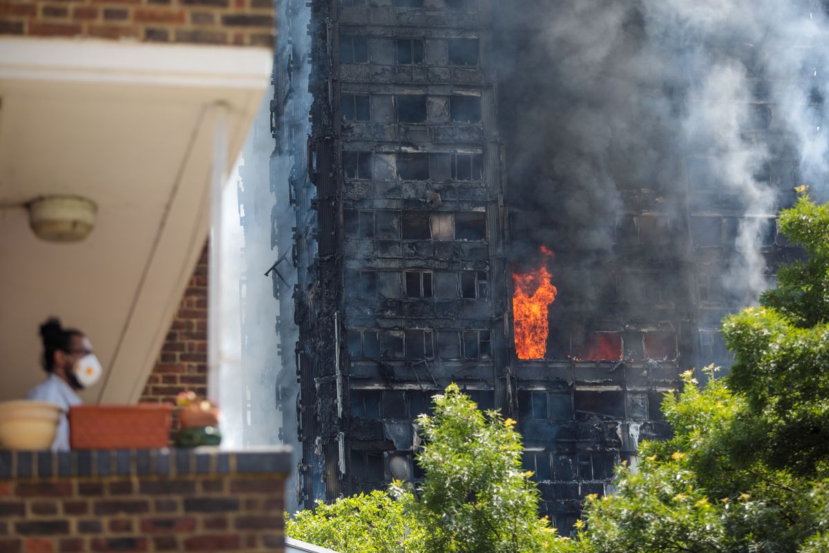 The building burns with flames coming out of windows in the background and a man wears a mask in the foreground. 
