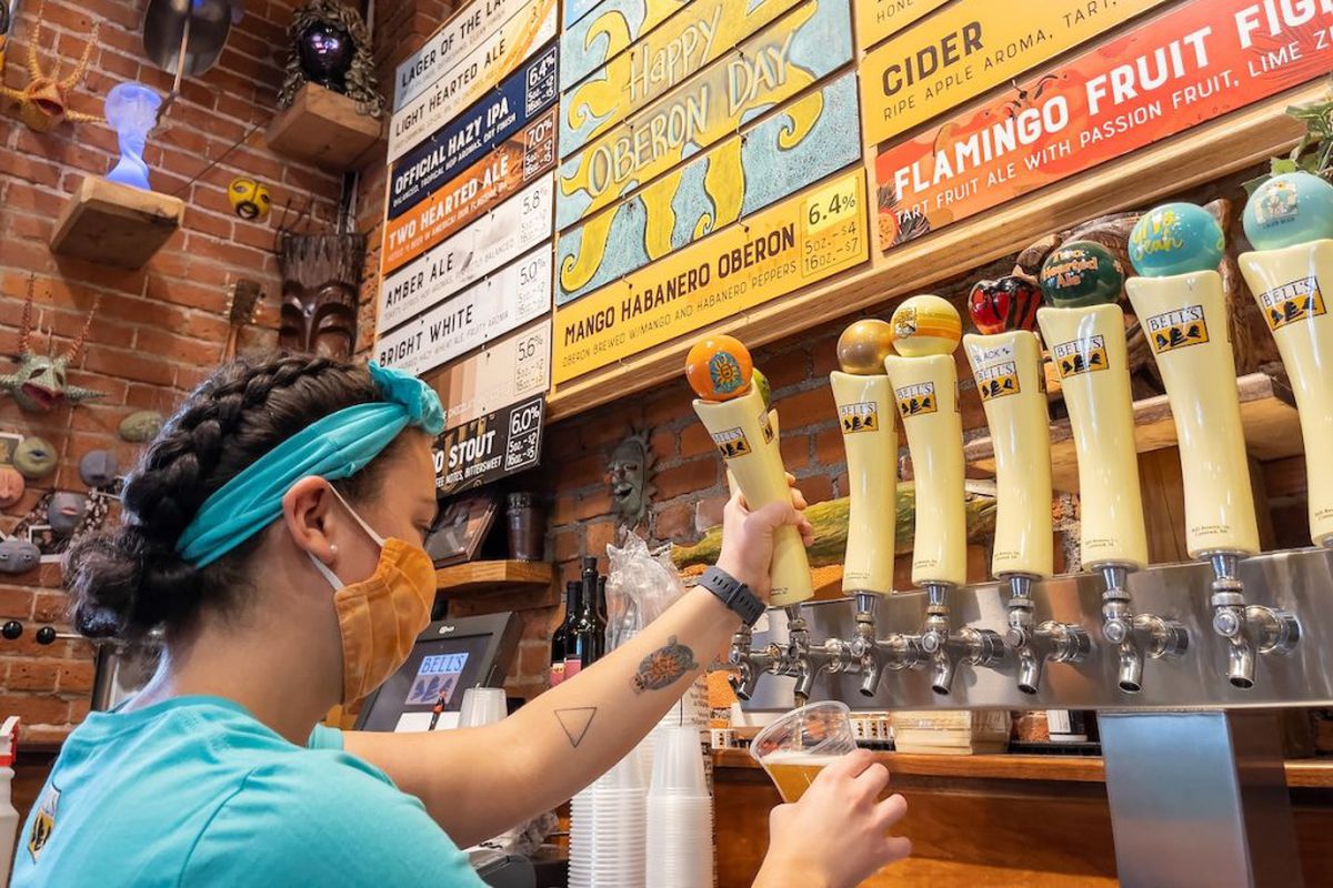 A woman wearing a mask pours a draft beer out of a branded Bell’s Brewing tapper.