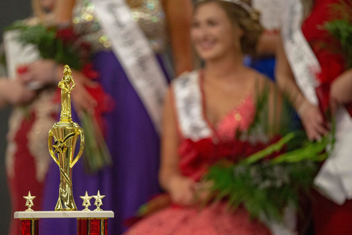 In the foreground, there is a tall, gold trophy with a red metallic base. In the distant background, out of focus, and large group of Beauty Queens sit as they prepare to be judged in the Miss Winnebago County Fair beauty pageant