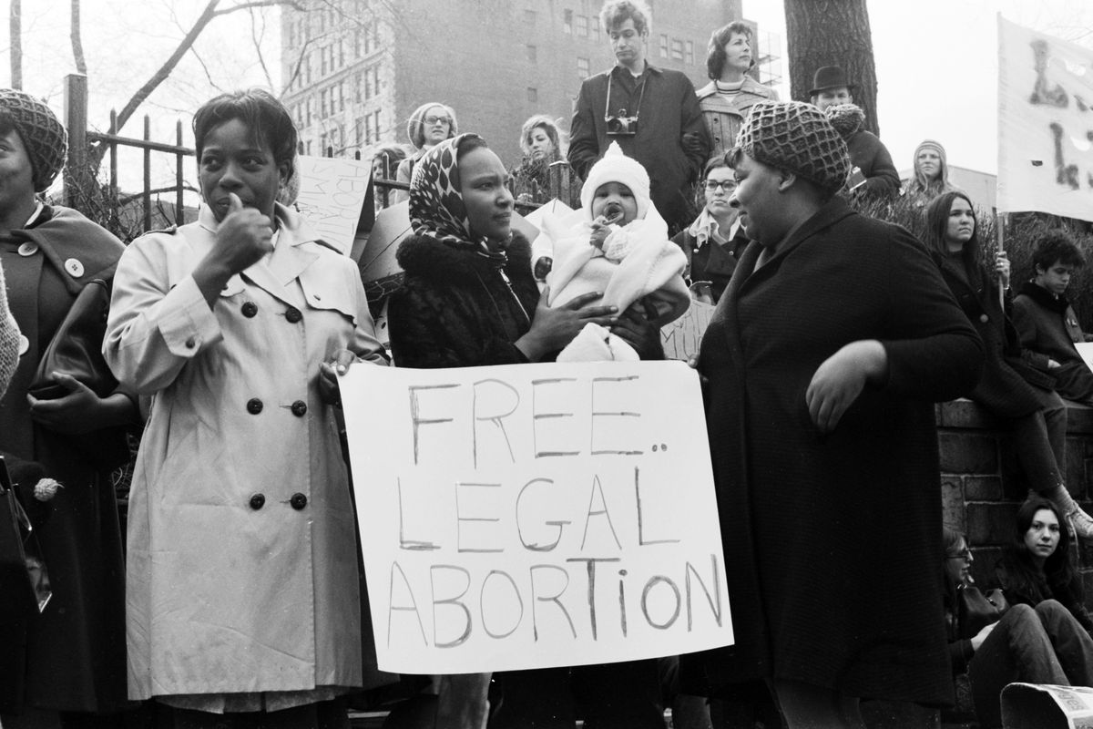 A group of women in hats and coats are led by several Black women holding a sign reading “Free Legal Abortion” on a city sidewalk.