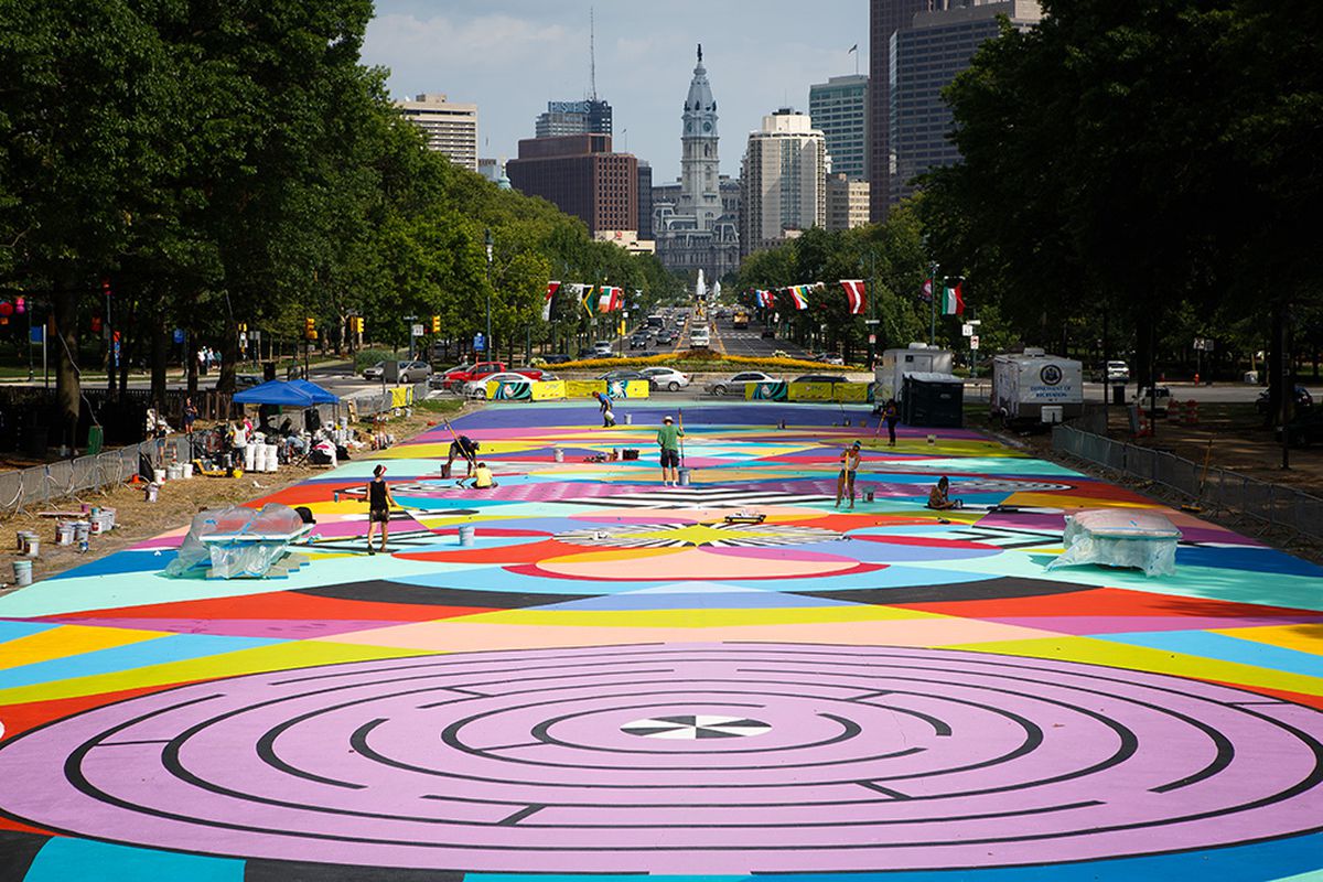 A colorful graphic mural is being painted in the street by people using giant brushes with the skyline of Philadelphia in the background.