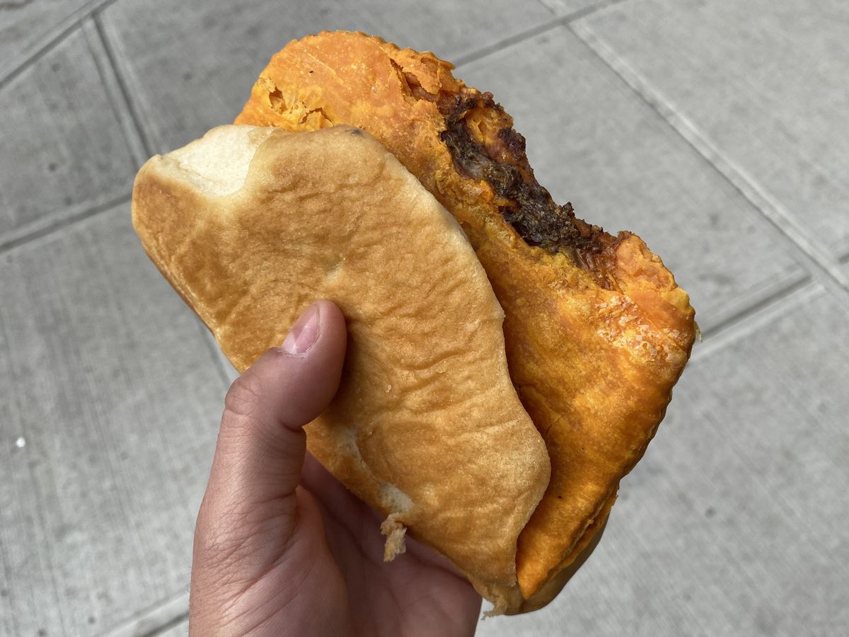 A hand clutches an orange beef patty with cheese wrapped in a piece of coco bread.