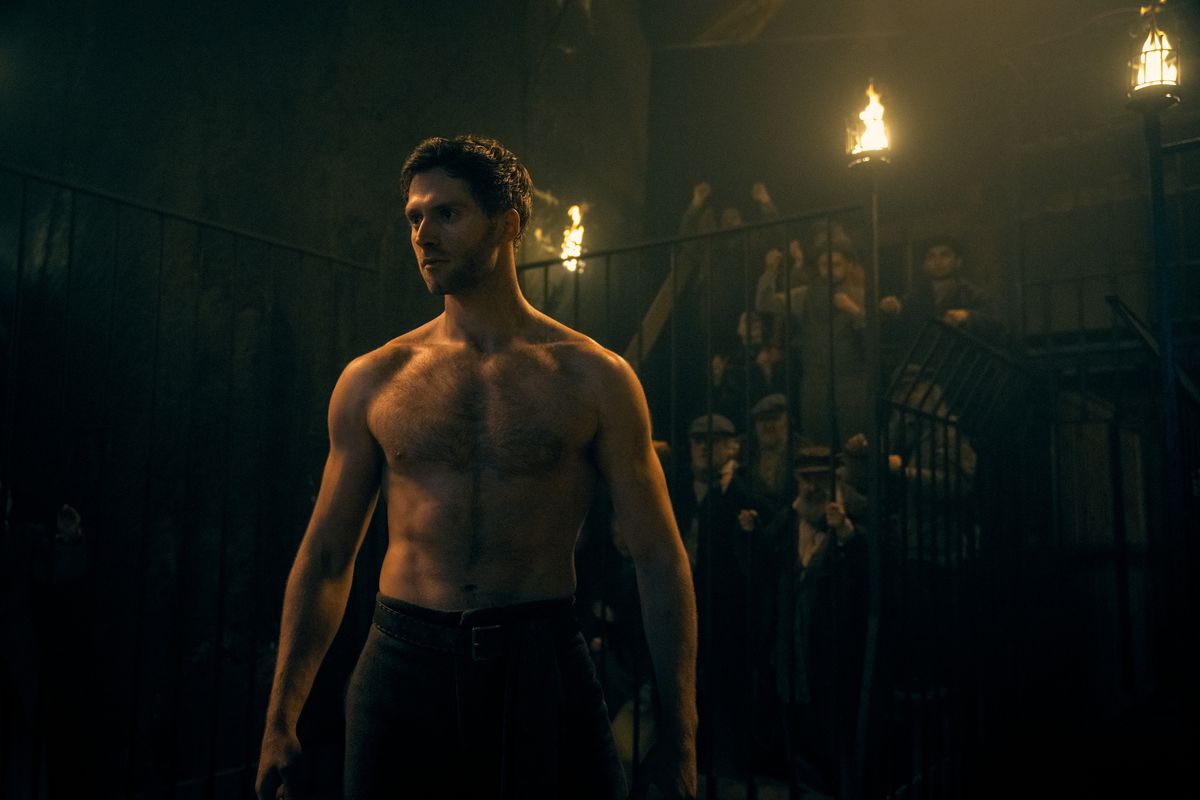 a shirtless, muscular man stands in what appears to be a fighting ring lit by torches