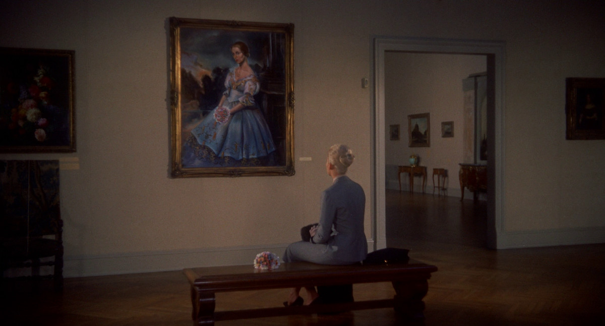 Kim Novak wears her iconic gray wool suit from Vertigo, while sitting at a museum bench and admiring a painting of a woman in a dress. Flowers adorn both women.