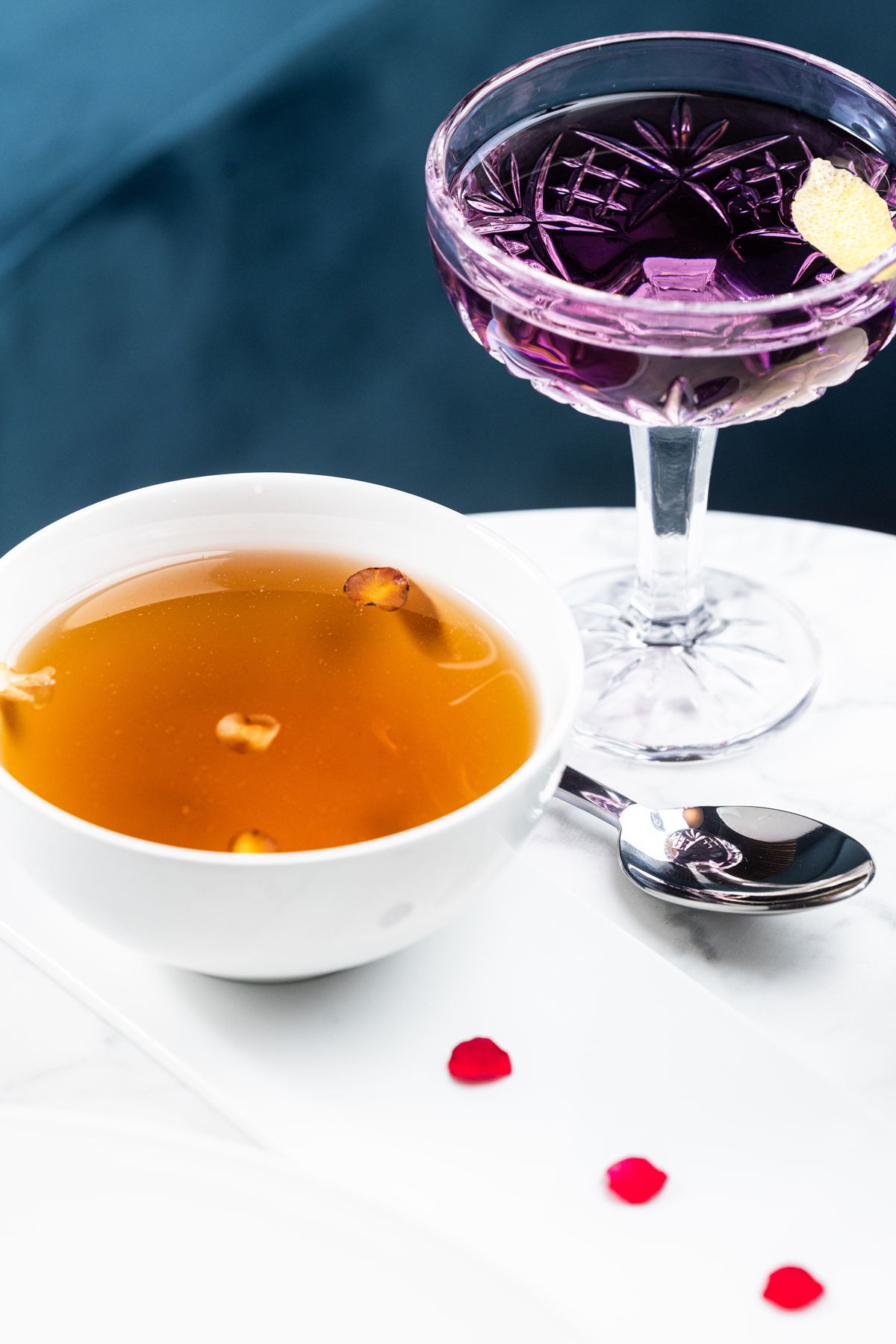 A white bowl filled with brown broth and a few floating flower petals is on the left. On the right is an engraved glass coupe with a purple cocktail and a twist of lemon.