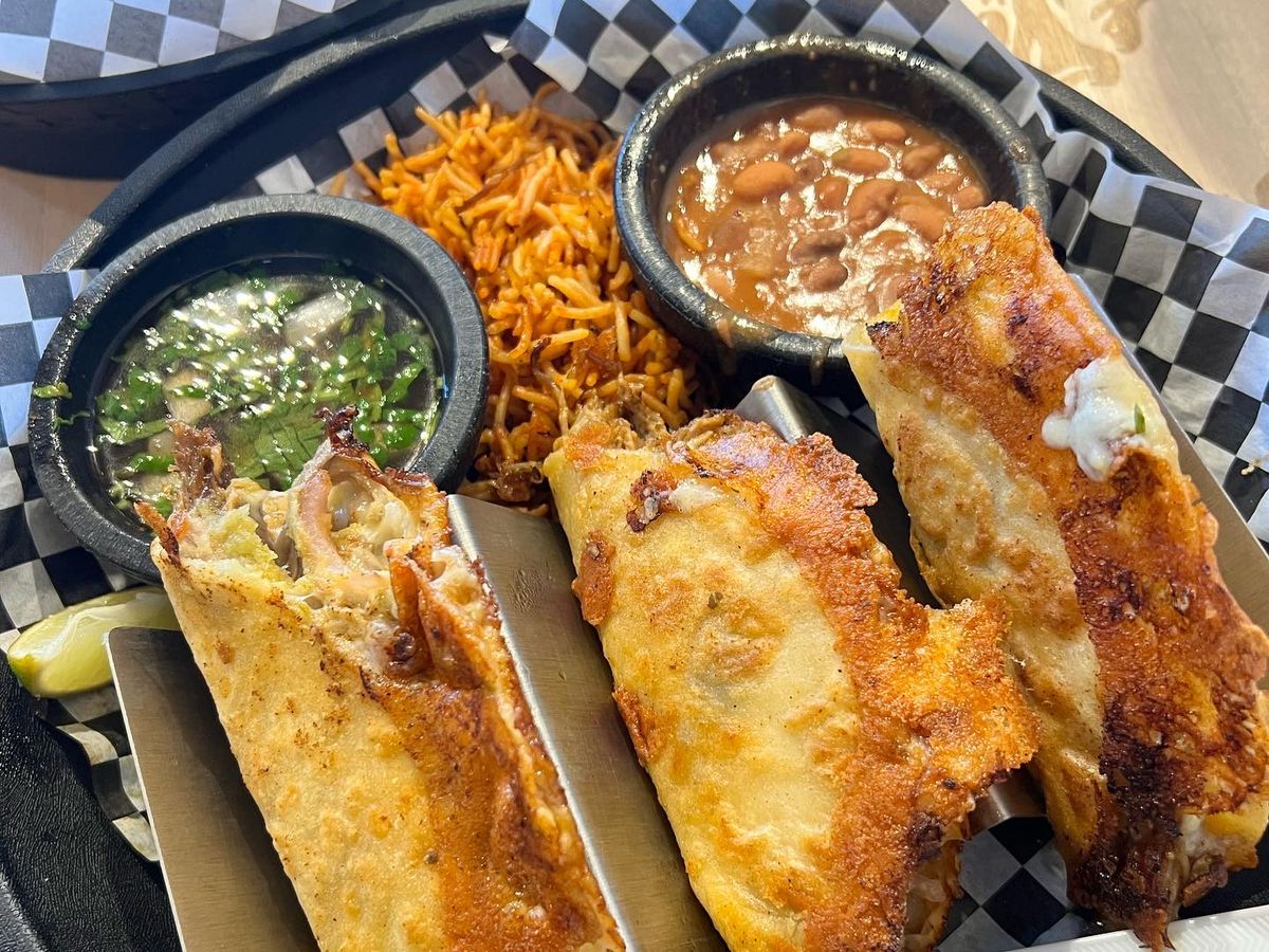 A plastic boat lined with checkerboard butcher paper. The boat holds three tacos, crusted with cheese, alongside a small pile of fideos, and cups of pinto beans and salsa