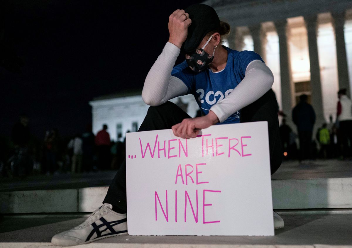 Woman wearing mask and 2020 T-shirt holds white sign saying “When There Are Nine” in pink marker. Her head is in her hands, and it appears as if she may be crying. She sits on the curb in front of the Court, a loose crowd visible behind her.