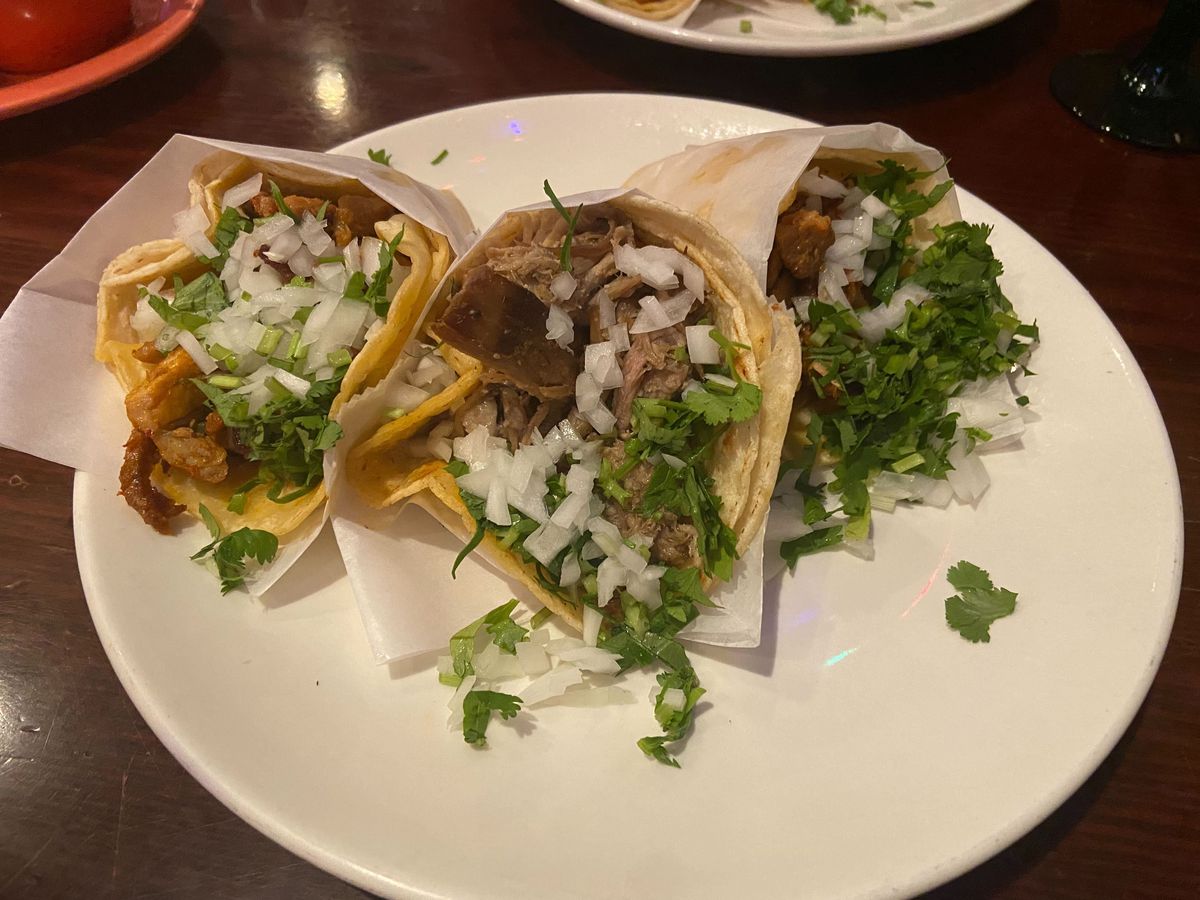 Three cone-shaped, meat-filled tacos are covered in white onion and cilantro with wax wrapping, sitting atop a white plate on a black table.