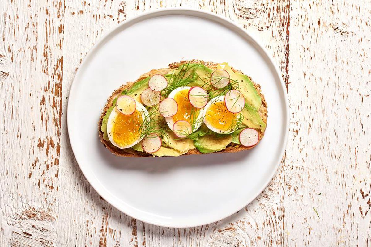 Overhead view of a piece of bread topped with avocado, sliced soft egg, and sliced radishes, on a white plate