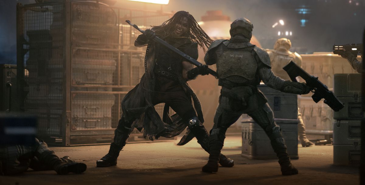 Bloodaxe (Ray Fisher), a man with small dreadlocks dressed in overlapping layers of leather and metal and wielding a metal staff, strikes an armored trooper in a warehouse-like space full of shelves, storage, and boxes in Netflix’s Rebel Moon