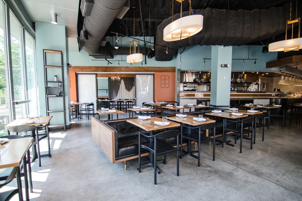 A wide shot of a restaurant space with a polished cement floor, light blue walls, light wooden tables with a mix of black chairs and low black banquette seating, and some orange accents.