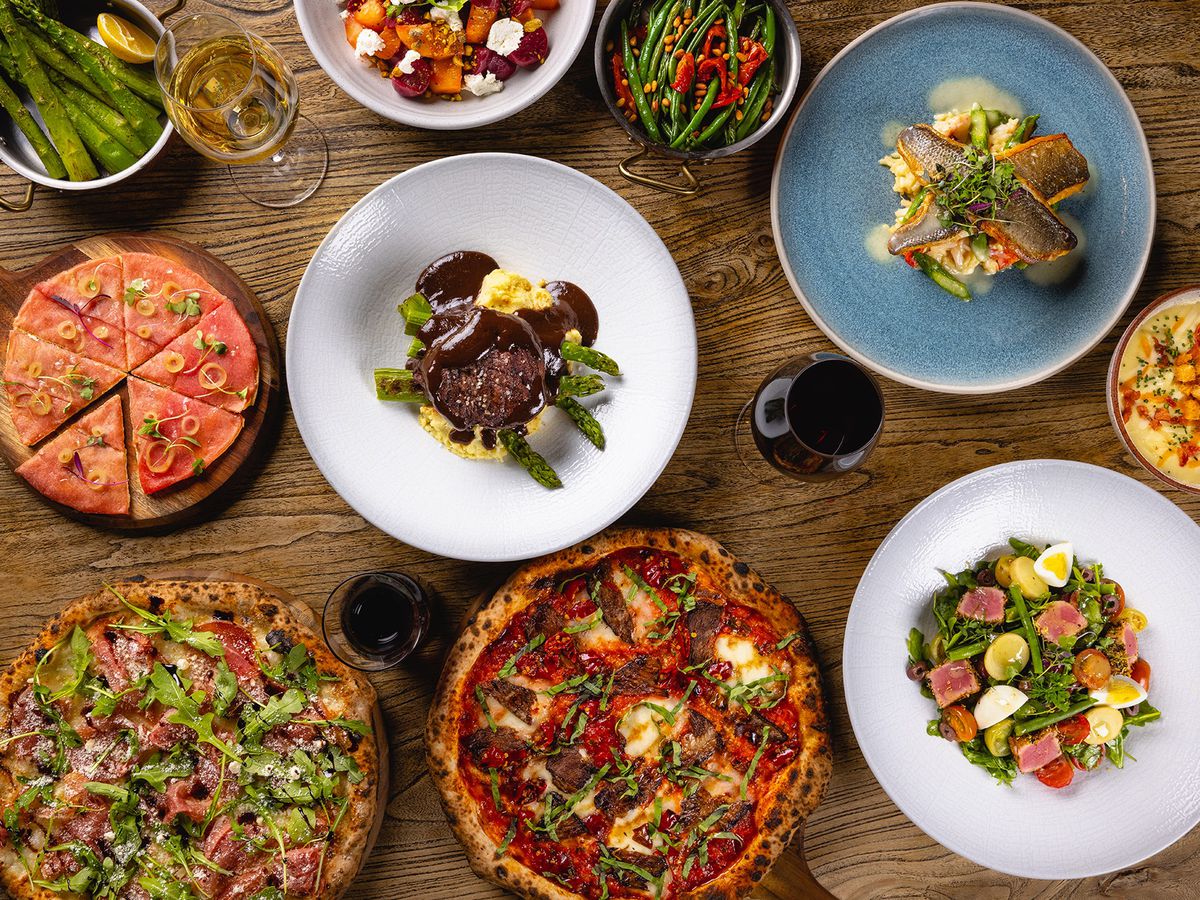 An overhead view of pizzas, a pasta dish, beef, and more on a wood table