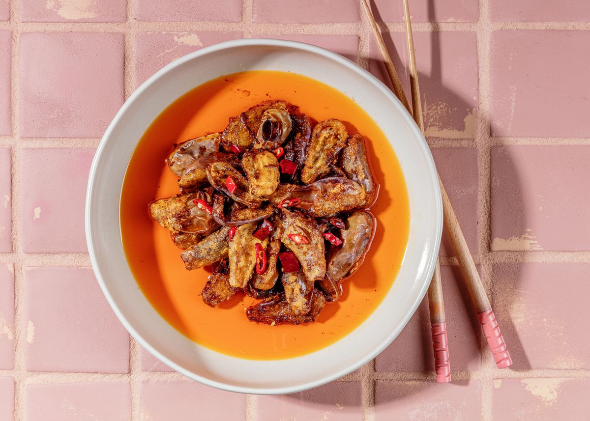 Tiny eggplants swimming in chili oil in a white bowl on pink tile with chopstick to the side featuring pink ends. 