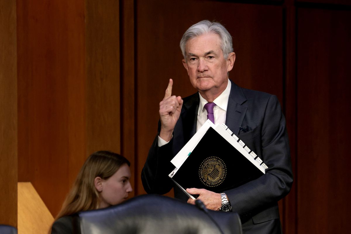 Powell, gray-haired and clean-shaven, in a dark suit, white shirt, and red tie, holds a folder with the Fed’s logo against his chest and points to the ceiling with his free hand.