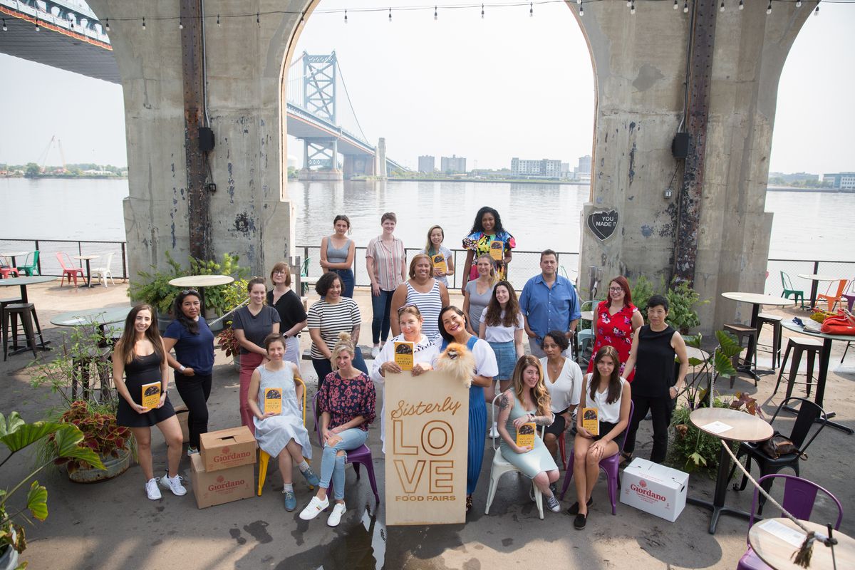 a group of women gather on a pier in front of a sign for Sisterly Love Food Fair